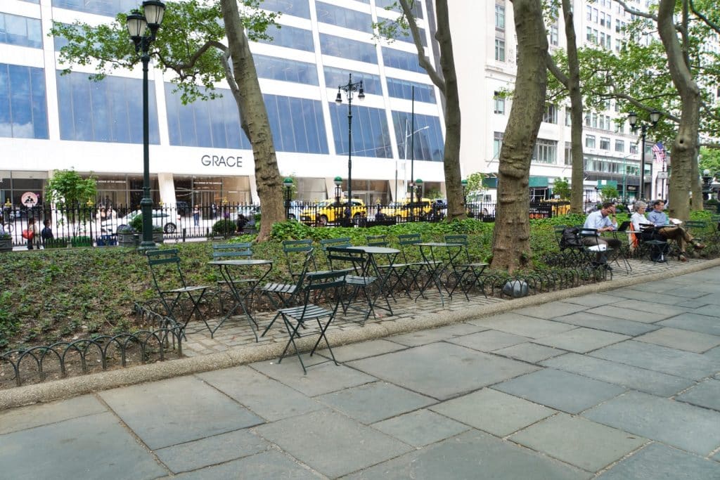 Grüne Klappstühle und Tische stehen auf einem gepflasterten Bereich im Bryant Park in New York, während im Hintergrund das Grace Building und Passanten zu sehen sind.