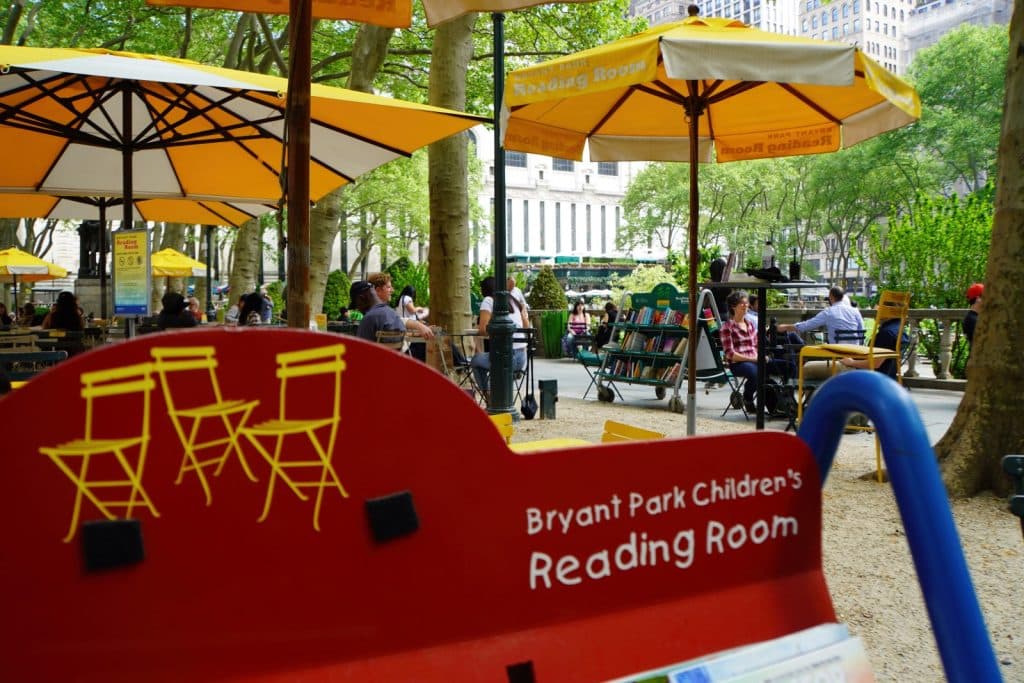 Der Bryant Park Children's Reading Room in New York zeigt einen Außenbereich mit gelben Sonnenschirmen, Sitzgelegenheiten und einem mobilen Bücherregal unter schattenspendenden Bäumen. Im Vordergrund ist ein rotes Schild mit gelben Stuhlgrafiken und der Aufschrift „Bryant Park Children's Reading Room“ zu sehen.
