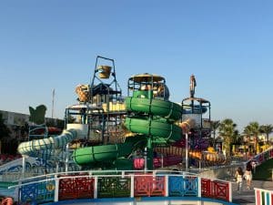 Ein farbenfroher Wasserpark im Rixos Premium Seagate mit mehreren verschlungenen Wasserrutschen, einem großen Kippeimer und einem hölzernen Brückengeländer im Vordergrund unter blauem Himmel.