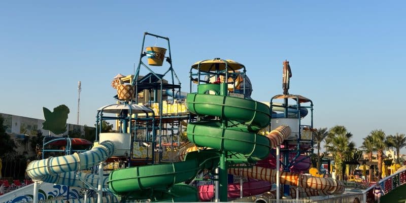 Ein farbenfroher Wasserpark im Rixos Premium Seagate mit mehreren verschlungenen Wasserrutschen, einem großen Kippeimer und einem hölzernen Brückengeländer im Vordergrund unter blauem Himmel.