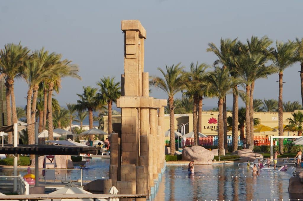 Ein weitläufiger Außenpool im Rixos Premium Seagate mit antiken ägyptischen Säulenrepliken im Wasser, umgeben von Palmen und einem Spa-Gebäude im Hintergrund.