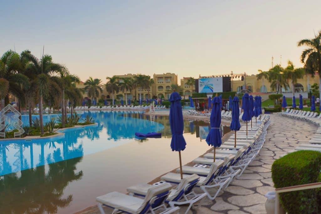Ein weitläufiger, geschwungener Außenpool im Rixos Sharm El Sheikh Resort, gesäumt von Palmen, weißen Sonnenliegen mit blauen Sonnenschirmen und Hotelgebäuden im Hintergrund bei Sonnenuntergang.
