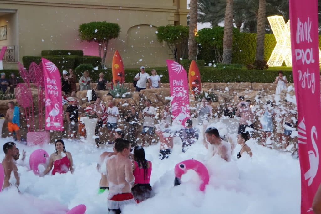Gäste feiern bei einer „Pink Pool Party“ im Rixos Sharm El Sheikh in einem mit weißem Schaum gefüllten Pool, dekoriert mit rosa Flamingos und passenden Bannern.