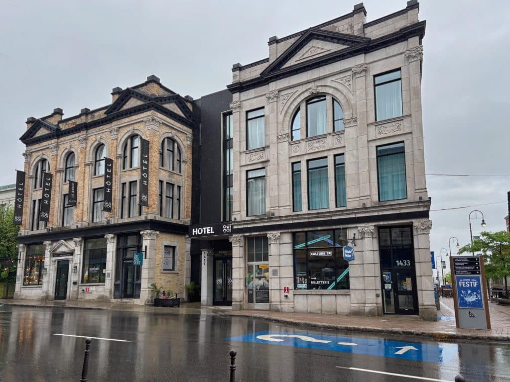 Zwei historische, mehrstöckige Steingebäude des Hotels Oui Go! in Trois-Rivières stehen an einer regennassen Straße unter bewölktem Himmel.