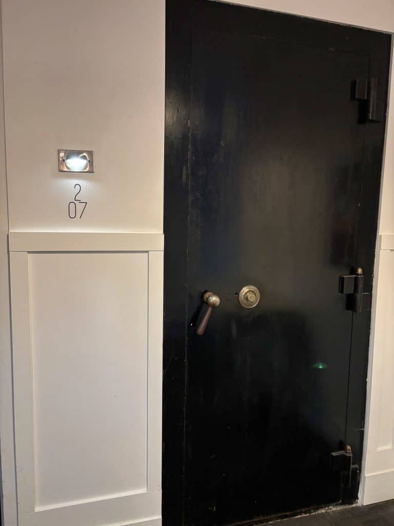 Eine massive, schwarze Tresortür mit großen Scharnieren und einem Drehgriff befindet sich in einem Hotel in Trois-Rivières direkt neben der weißen Wand von Zimmer 207.