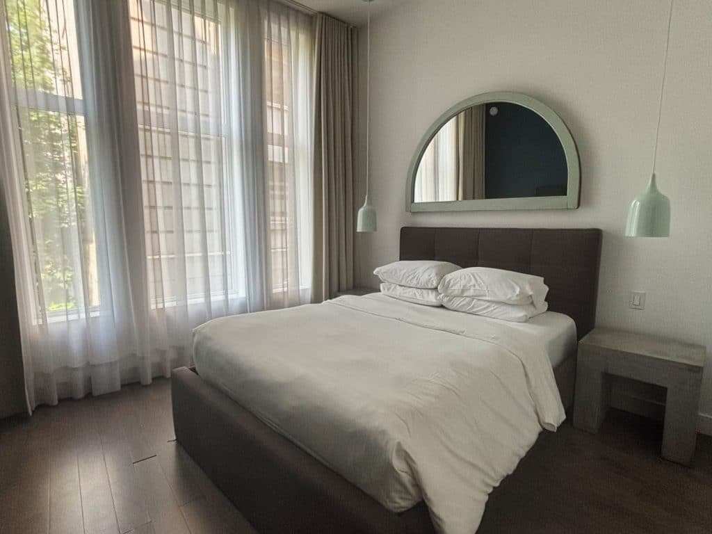 Ein modernes Hotelzimmer in Trois-Rivières mit einem großen Bett mit weißer Bettwäsche, einem bogenförmigen Spiegel über dem gepolsterten Kopfteil und bodentiefen Fenstern mit hellen Vorhängen.