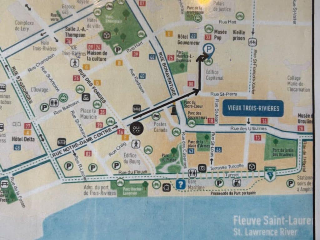 Ein Ausschnitt eines Stadt- und Parkplans von Trois-Rivières zeigt Straßenverläufe, Sehenswürdigkeiten wie das Hotel Gouverneur und Grünflächen, wobei eine schwarze Pfeillinie eine Route von der Rue Notre-Dame Centre zu einem Parkplatz markiert.