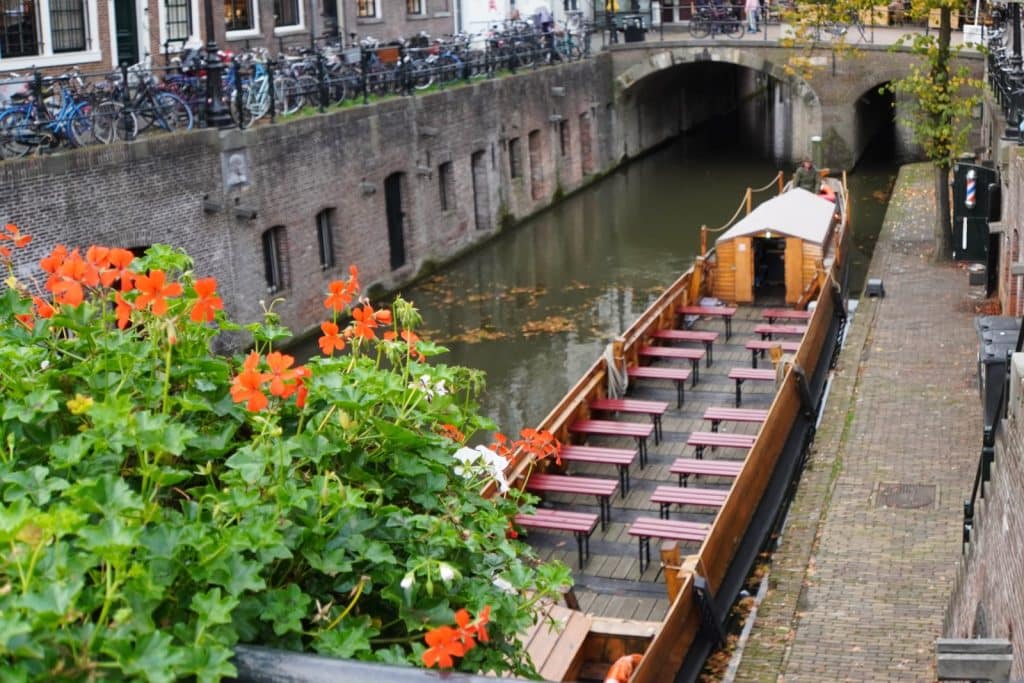 Ein hölzernes Boot, gestaltet als römisches Schiff mit zahlreichen Sitzbänken, liegt in einer Gracht in Utrecht unterhalb einer historischen Steinbrücke. Im Vordergrund blühen leuchtend orangefarbene Geranien, während am gegenüberliegenden Ufer zahlreiche Fahrräder an einem Geländer lehnen.