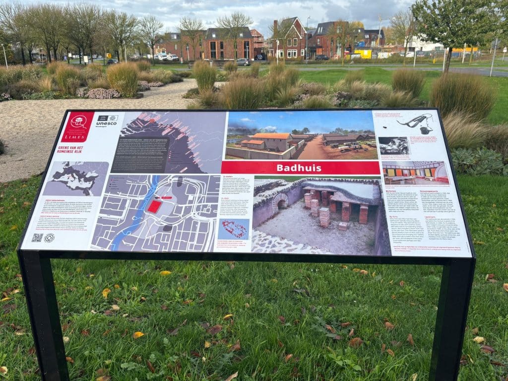 Eine Informationstafel im Freien zeigt Illustrationen, Karten und Texte zum römischen Badehaus des Castellums in Utrecht. Die Tafel präsentiert eine Rekonstruktionszeichnung des Gebäudes sowie ein Foto der archäologischen Ausgrabungen mit den Überresten der Hypokaustum-Heizung.