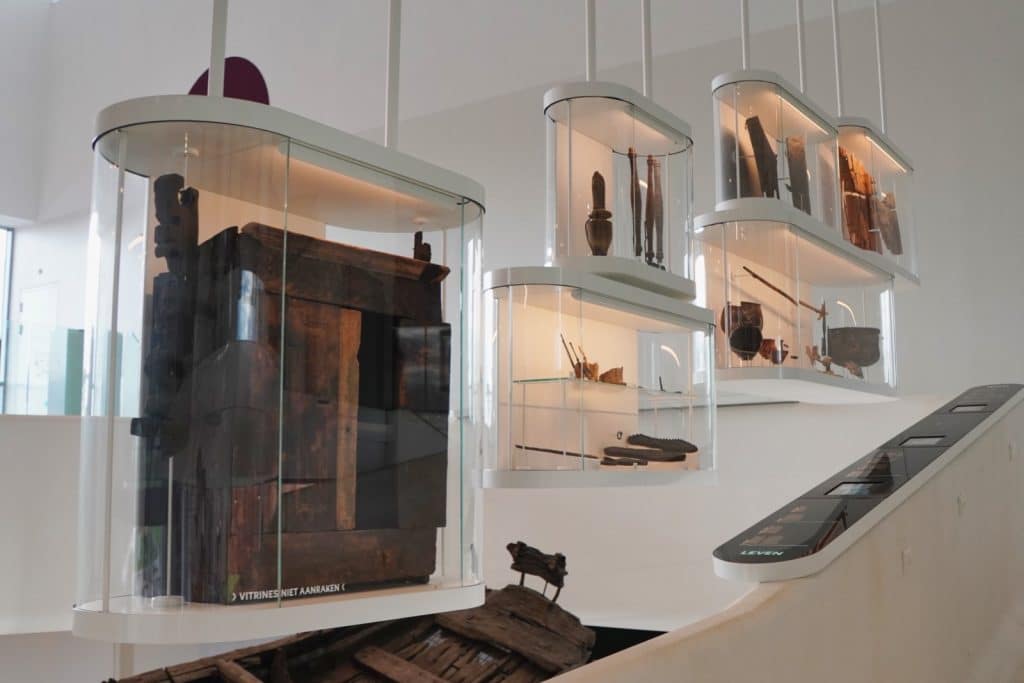 In einem modernen Ausstellungsraum des Museum Hoge Woerd in Utrecht hängen mehrere beleuchtete Glasvitrinen mit archäologischen Funden über den hölzernen Überresten eines antiken römischen Schiffes.