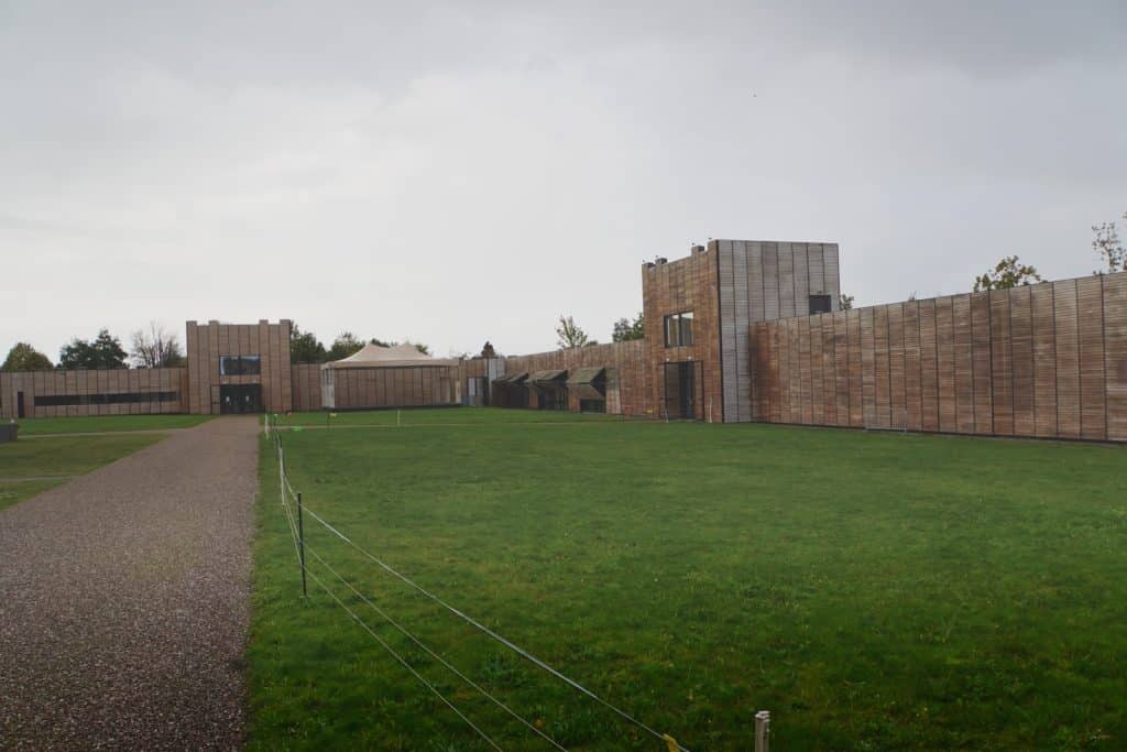 Der Innenbereich des Castellum Hoge Woerd in Utrecht zeigt eine weite Rasenfläche, die von modernen, hohen Holzmauern und Wachtürmen umschlossen ist, mit einem Kiesweg im Vordergrund.