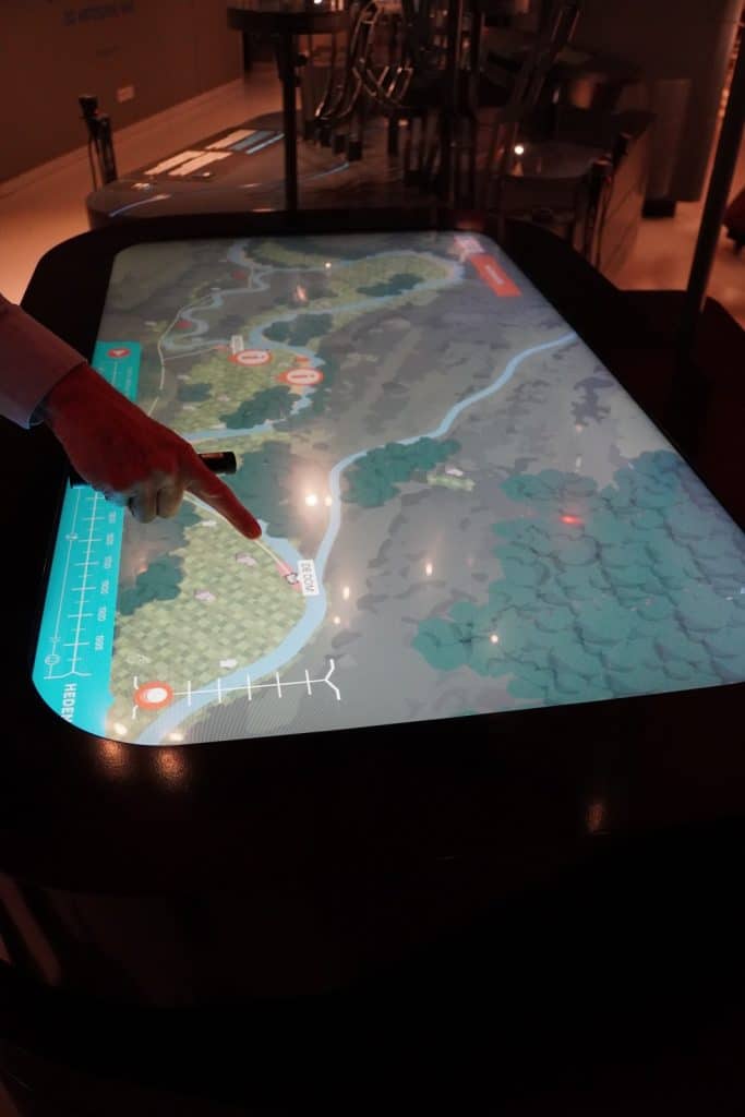 Eine Person deutet mit dem Finger auf eine interaktive, leuchtende Karte, die auf einem großen Touch-Tisch in einem abgedunkelten Ausstellungsraum präsentiert wird. Die digitale Karte zeigt eine grüne Landschaft mit Flussläufen und verschiedenen Markierungspunkten zur historischen Erkundung.