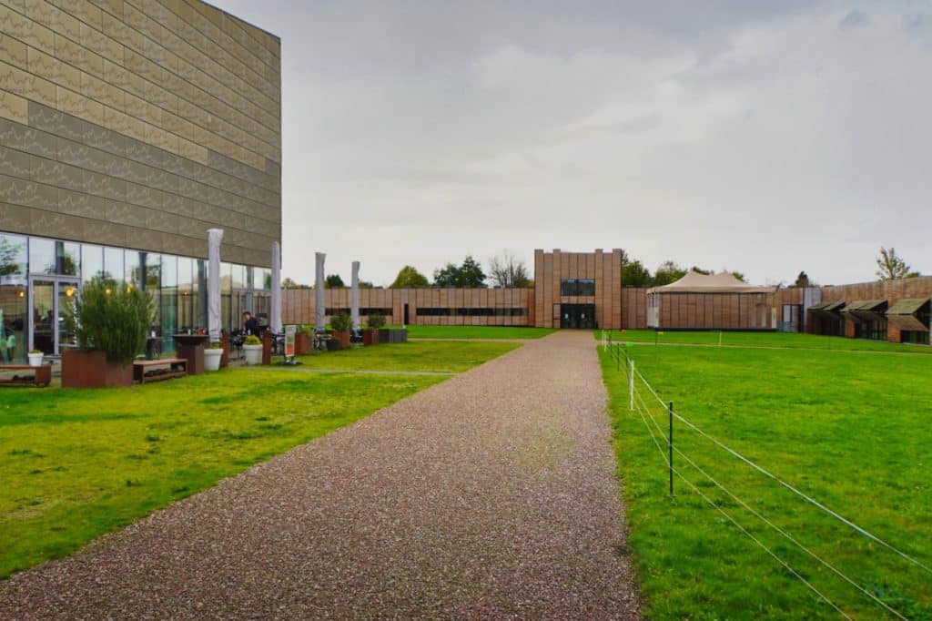 Ein breiter Kiesweg führt über eine grüne Wiese auf den modernen Nachbau des römischen Castellum Hoge Woerd in Utrecht zu, dessen hölzerne Fassaden und Glasfronten ein Café mit Außenbereich beherbergen.