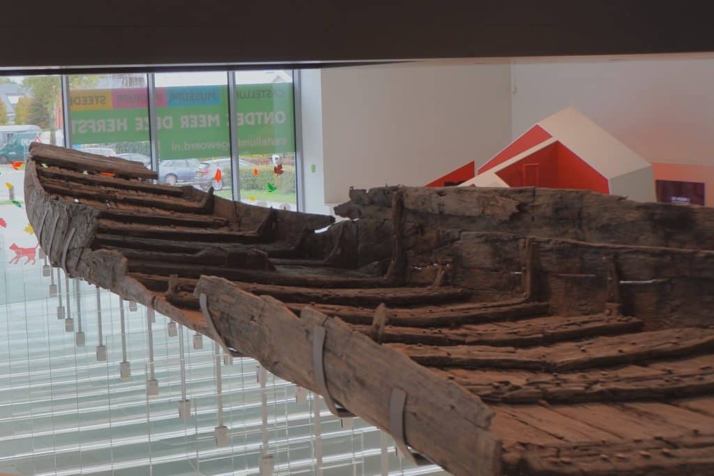 Das antike römische Holzschiff „De Meern 1“ ist im Museum Castellum Hoge Woerd in Utrecht ausgestellt und wird von einer modernen Metallkonstruktion über einem hellen Ausstellungsraum getragen.