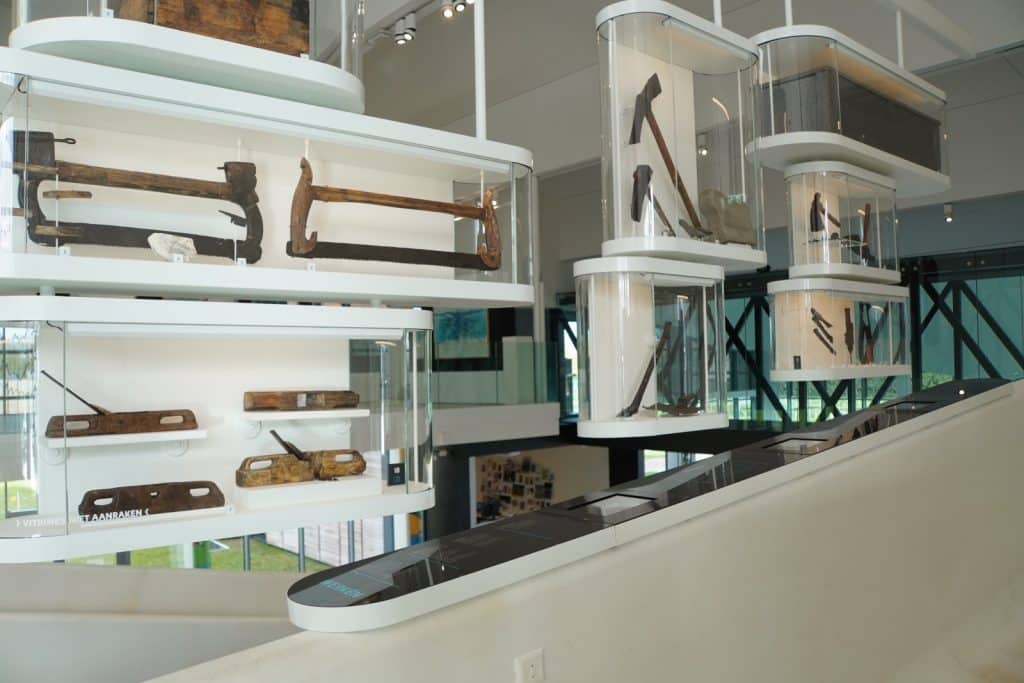 In einem modernen Ausstellungsraum im Museum Hoge Woerd in Utrecht werden seltene historische Werkzeuge, wie Sägen, Äxte und Hobel, in schwebenden, weiß gerahmten Glasvitrinen präsentiert. Die Vitrinen sind versetzt übereinander angeordnet und bieten einen Blick auf die archäologischen Fundstücke vor einem hellen, minimalistischen Hintergrund.