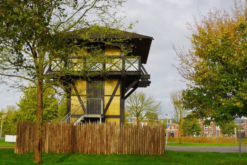 Rekonstruktion eines römischen Wachturms am Castellum Utrecht mit gelben Wänden, dunklem Fachwerk und einem hölzernen Palisadenzaun im Vordergrund.