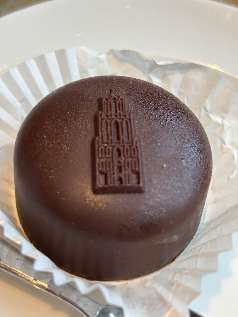 Ein rundes, mit dunkler Schokolade überzogenes Törtchen aus dem Dom Café in Utrecht, das auf der Oberseite ein Relief des Utrechter Domturms zeigt. Es liegt in einer weißen Papierform auf einem hellen Teller.