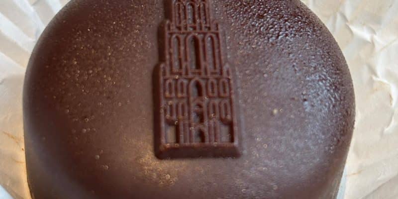 Ein rundes, mit dunkler Schokolade überzogenes Törtchen aus dem Dom Café in Utrecht, das auf der Oberseite ein Relief des Utrechter Domturms zeigt. Es liegt in einer weißen Papierform auf einem hellen Teller.