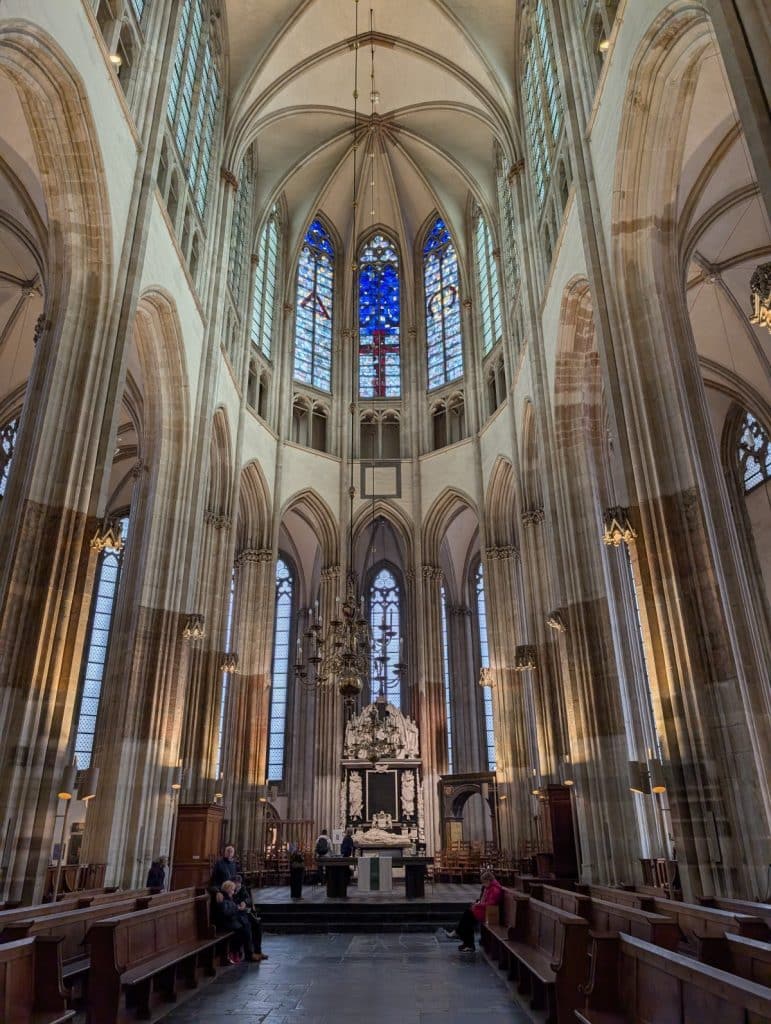 Blick in den hoch aufragenden gotischen Chor des Utrechter Doms mit seinen schlanken Bündelpfeilern, spitzzulaufenden Gewölben und farbenprächtigen Glasfenstern über dem Altarraum. Im Vordergrund sind hölzerne Kirchenbänke und einige Besucher in der weitläufigen Architektur zu sehen.