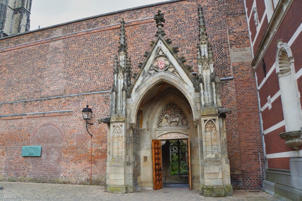Ein gotisches Sandsteinportal mit kunstvollen Schnitzereien und einem Wappen führt in den Garten des Utrechter Doms, flankiert von einer hohen Backsteinmauer und einer historischen Straßenlaterne.