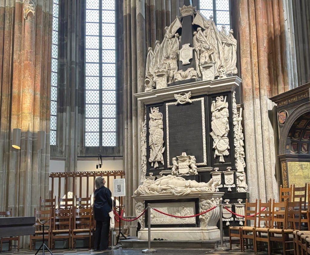 Ein monumentales, barockes Grabmal im Utrechter Dom mit einer liegenden Marmorfigur auf einem Sarkophag, flankiert von hohen Säulen und aufwendigen Skulpturen. Eine Person steht davor und betrachtet die imposante Gedenkstätte, die vor hohen gotischen Fenstern platziert ist.