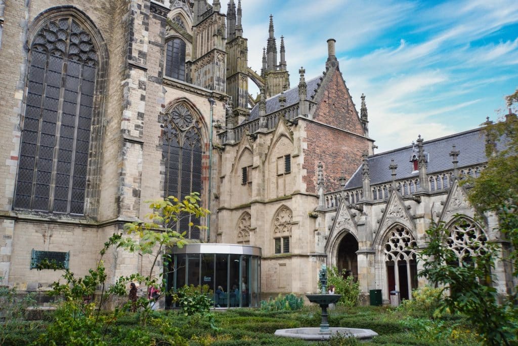 Der Innenhof des Utrechter Doms zeigt ein modernes Glascafé inmitten historischer gotischer Architektur mit Spitzbogenfenstern, einem Springbrunnen und gepflegten Grünanlagen.