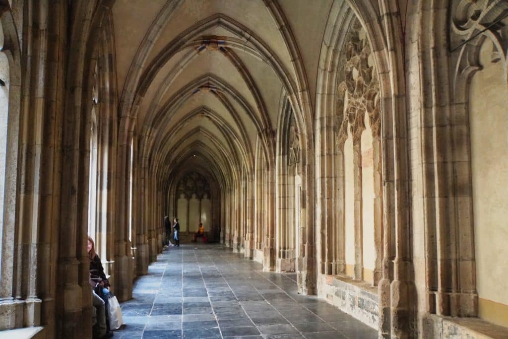 Ein langer, steinerner Gang im Kreuzgang des Utrechter Doms mit hohen gotischen Spitzbogengewölben, kunstvollen Maßwerkfenstern und einigen Personen, die auf den seitlichen Steinbänken verweilen.