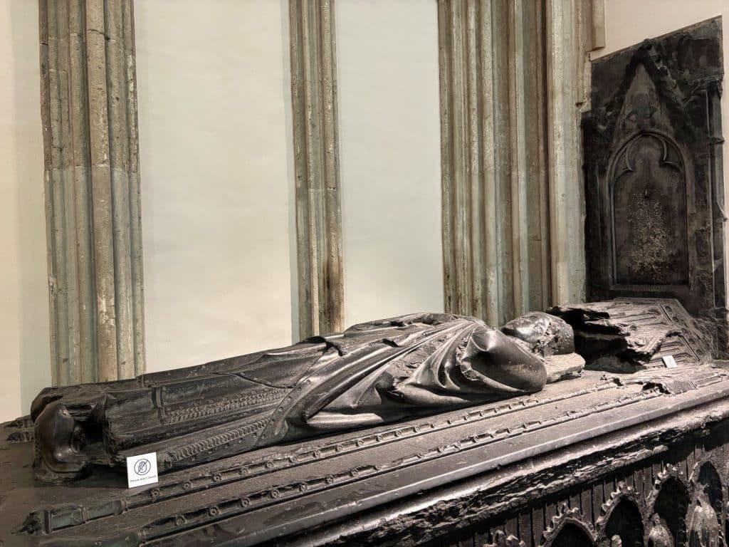 Ein detailreicher, steinerner Sarkophag im Utrechter Dom zeigt die liegende Skulptur einer verstorbenen Person in fließenden Gewändern vor einer Wand mit gotischen Säulenelementen.