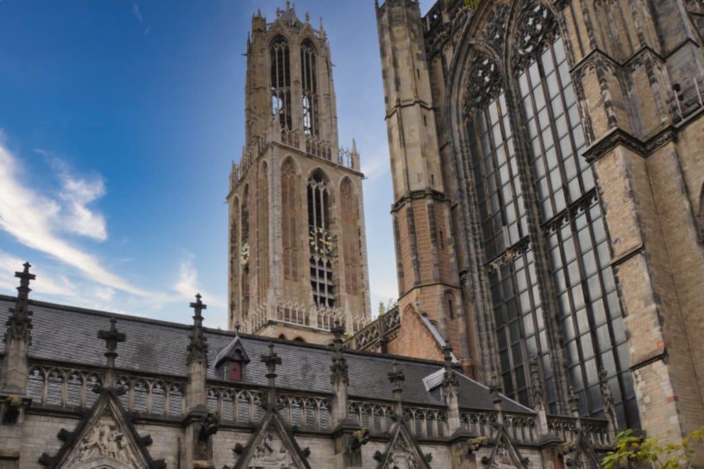 Blick aus dem Garten auf den historischen Domturm in Utrecht, der hinter den gotischen Mauern und großen Fenstern der Kathedrale in den blauen Himmel ragt.