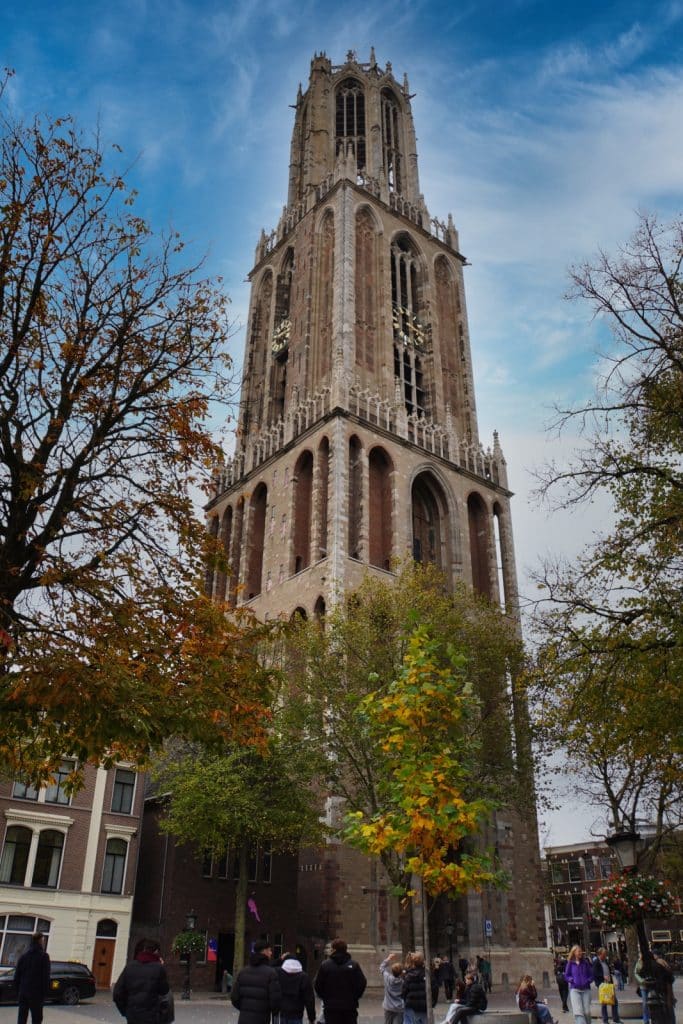 Der gotische Domturm von Utrecht ragt majestätisch hinter herbstlich gefärbten Bäumen in den blauen Himmel, während Passanten über den davorliegenden Platz spazieren.