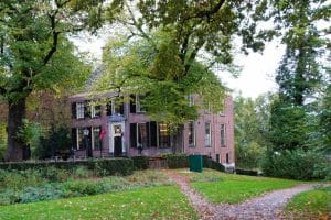 Ein zweistöckiges, historisches Backsteingebäude des Stayokay Hostel Bunnik mit dunklen Fensterläden, umgeben von herbstlichen Bäumen und einer gepflegten Grünanlage mit einem geschwungenen Kiesweg.