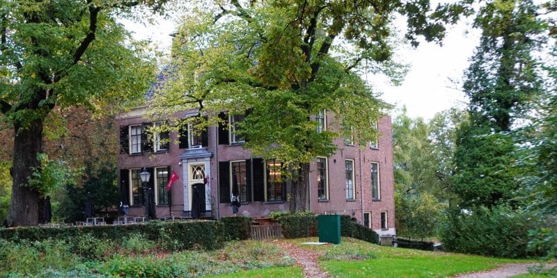 Ein zweistöckiges, historisches Backsteingebäude des Stayokay Hostel Bunnik mit dunklen Fensterläden, umgeben von herbstlichen Bäumen und einer gepflegten Grünanlage mit einem geschwungenen Kiesweg.