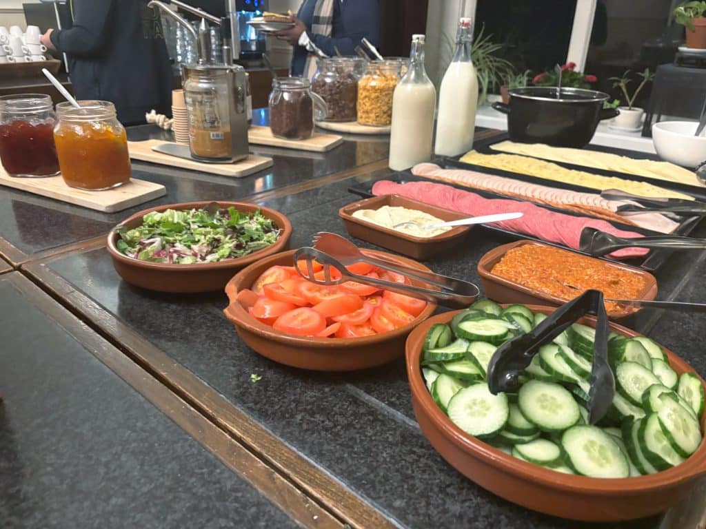 Ein reichhaltiges Frühstücksbuffet in einem Hostel bei Utrecht mit Schalen voller Gurkenscheiben, Tomaten und Salat sowie verschiedenen Aufstrichen, Wurst- und Käseplatten, Marmeladengläsern und Milchflaschen auf einer dunklen Theke.