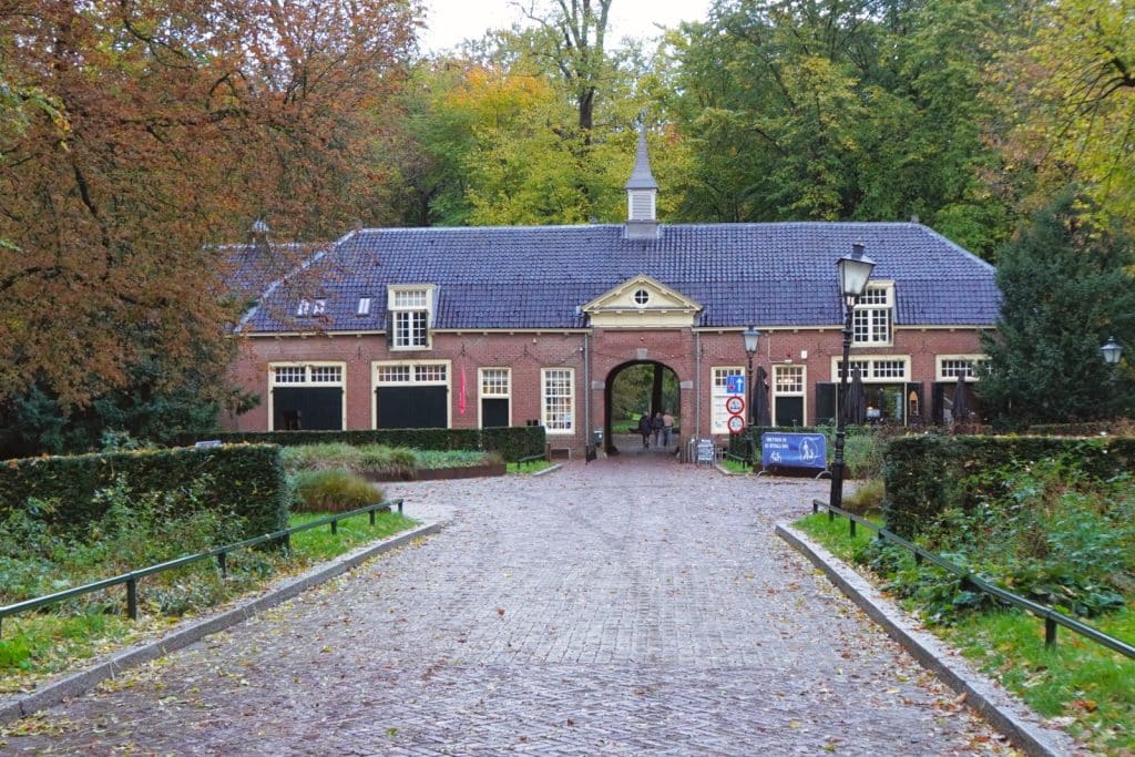 Ein gepflasterter Weg führt auf das historische, langgestreckte Backsteingebäude des Stayokay Hostel Bunnik zu, das durch ein zentrales Torhaus mit kleinem Turm und umliegende herbstliche Bäume geprägt ist.
