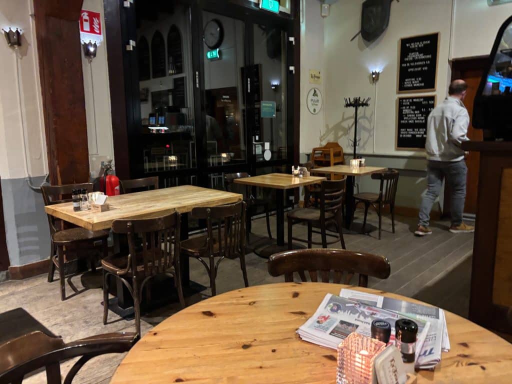 Innenansicht der gemütlichen Kneipe eines Hostels mit rustikalen Holztischen, Stühlen und einem Gast, der im Hintergrund an einer Menütafel vorbeiläuft.