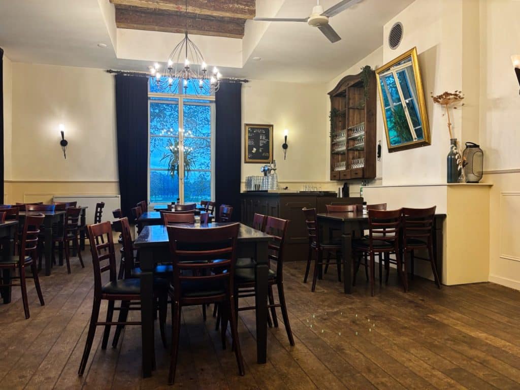 Ein gemütlicher Gastraum im Stayokay Hostel Bunnik mit dunklen Holztischen und Stühlen, einem großen Fenster mit blauen Vorhängen und einem eleganten Kronleuchter unter einer rustikalen Holzbalkendecke.