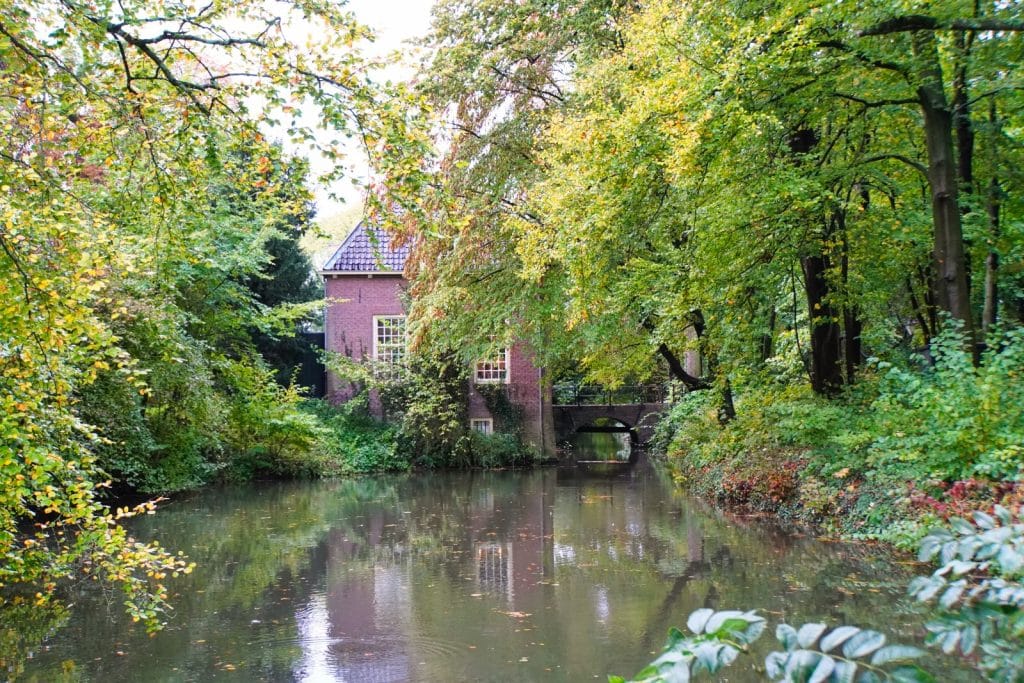 Ein idyllischer Blick auf das Stayokay Hostel Bunnik, ein Backsteingebäude am Ufer eines ruhigen Kanals, umgeben von dichten, herbstlich gefärbten Bäumen, die sich im Wasser spiegeln.