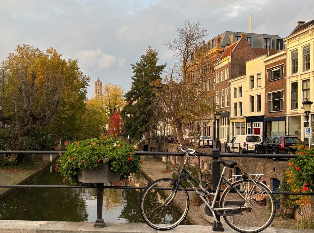Ein Fahrrad lehnt an einem Brückengeländer über einem Kanal in Utrecht, gesäumt von herbstlichen Bäumen, historischen Stadthäusern und dem im Hintergrund aufragenden Domturm.