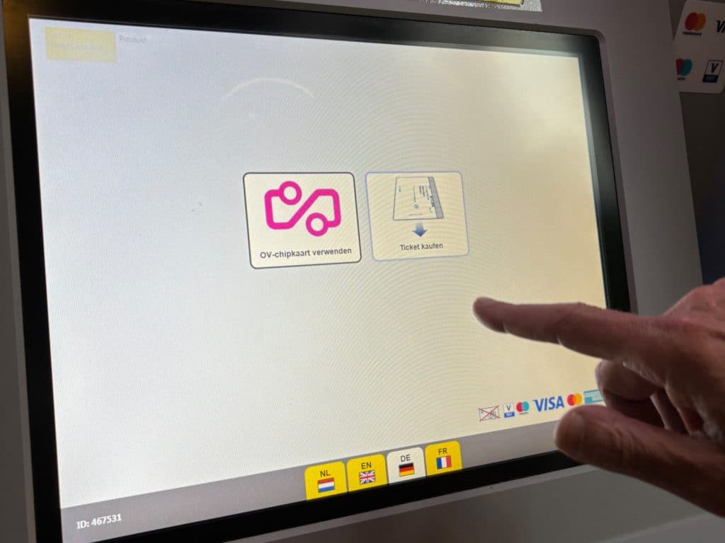 Ein Finger tippt auf den Touchscreen eines Parkscheinautomaten in Utrecht, der die Optionen „OV-chipkaart verwenden“ und „Ticket kaufen“ in deutscher Sprache anzeigt. Am unteren Bildschirmrand sind verschiedene Flaggen zur Sprachauswahl sowie Logos gängiger Zahlungsmittel wie Visa und Mastercard zu sehen.