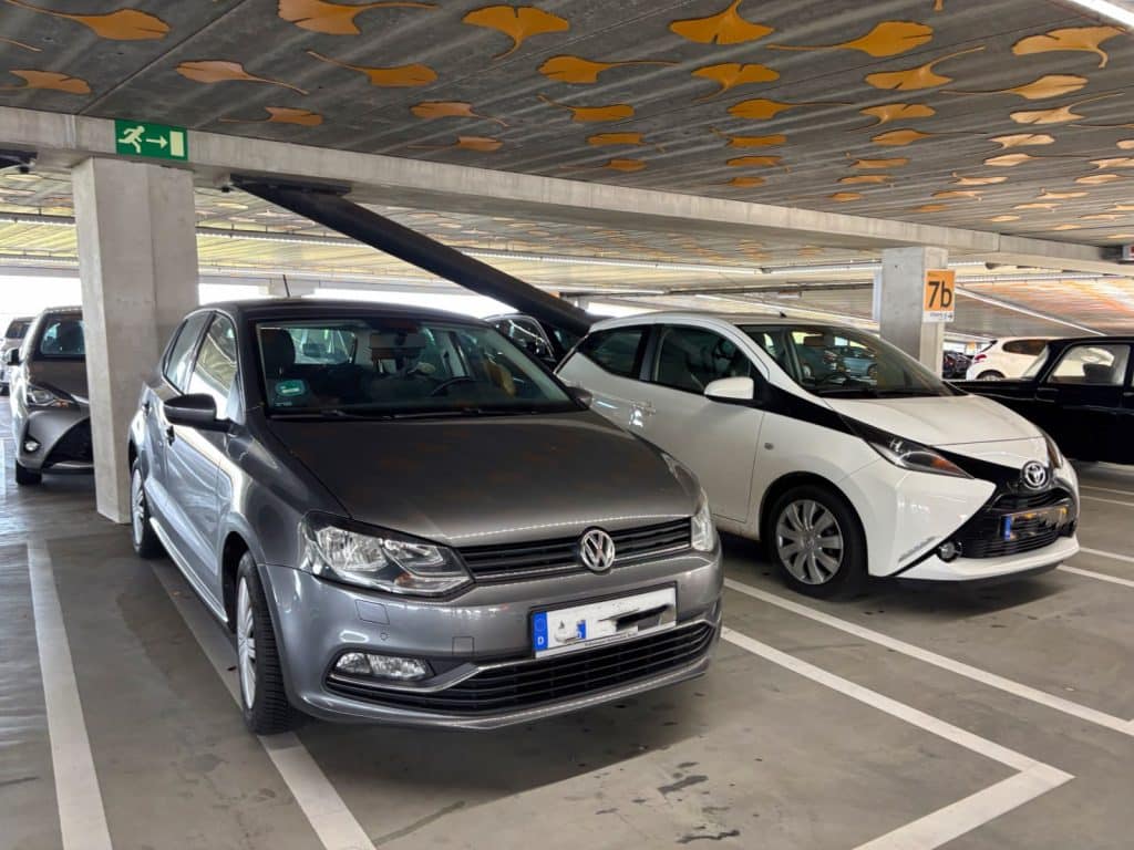 Ein grauer Volkswagen und ein weißer Toyota parken nebeneinander in einem Parkhaus in Utrecht, dessen Decke mit gelben Ginkgoblatt-Mustern verziert ist.
