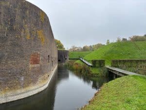 Eine massive, bogenförmige Backsteinmauer eines Festungsbauwerks der Holländischen Wasserlinie in Utrecht grenzt direkt an einen wassergefüllten Graben, neben dem ein hölzerner Steg an begrünten Erdwällen entlangführt.