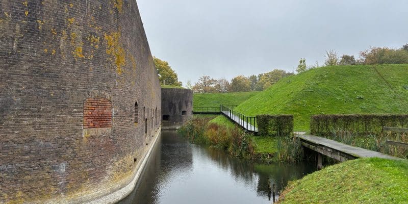 Eine massive, bogenförmige Backsteinmauer eines Festungsbauwerks der Holländischen Wasserlinie in Utrecht grenzt direkt an einen wassergefüllten Graben, neben dem ein hölzerner Steg an begrünten Erdwällen entlangführt.