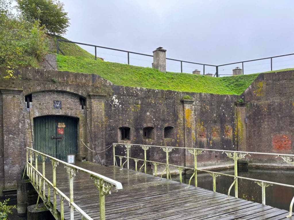 Eine hölzerne Brücke mit weißem Metallgeländer führt über einen Wassergraben zu einem massiven Backsteingebäude der Utrechter Wasserlinie, das eine grüne Holztür und grasbewachsene Wälle besitzt.