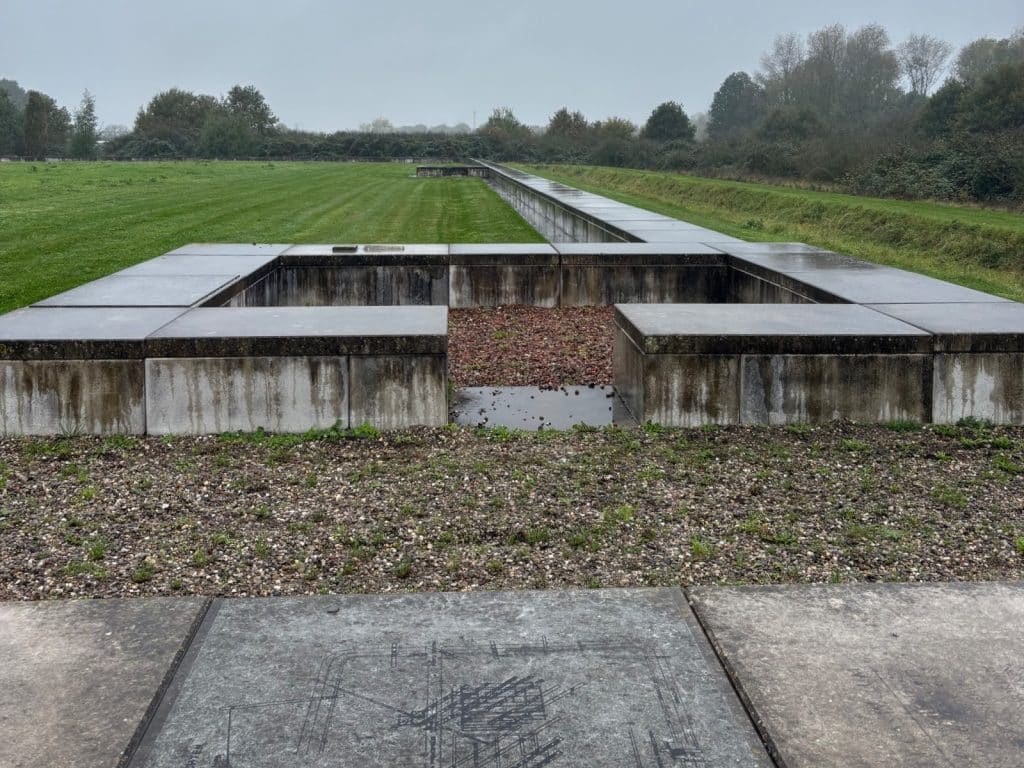 Eine weitläufige Außenanlage eines Castellum der Utrechter Wasserlinie zeigt flache, graue Betonstrukturen, die die Grundrisse eines ehemaligen römischen Kastells auf einer grünen Wiese markieren. Im Vordergrund ist eine Steinplatte mit einer eingravierten technischen Zeichnung zu sehen, während sich im Hintergrund eine lange Betonmauer unter einem bewölkten Himmel erstreckt.