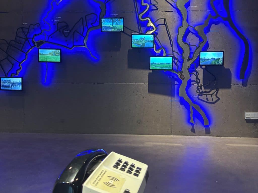 Eine interaktive Wandinstallation im Waterline Museum Utrecht zeigt eine beleuchtete Karte mit blau hinterlegten Wasserwegen und mehreren kleinen Bildschirmen, die Landschaften präsentieren, während im Vordergrund ein Audioguide-Telefon mit Tastenfeld zu sehen ist.