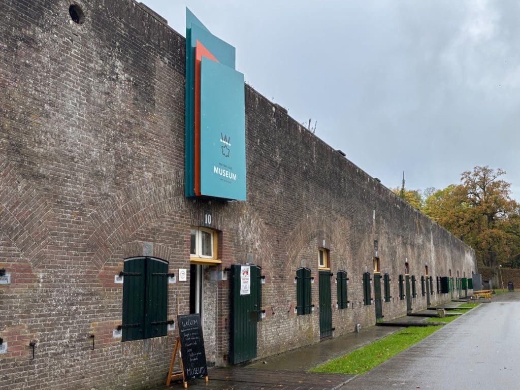 Der Eingang des Waterline Museums in Utrecht befindet sich in einem langgestreckten, historischen Backsteingebäude mit zahlreichen Rundbogennischen und grünen Fensterläden. Ein modernes, blau-oranges Schild mit dem Museumslogo ragt über dem Eingangsbereich an der Fassade empor.