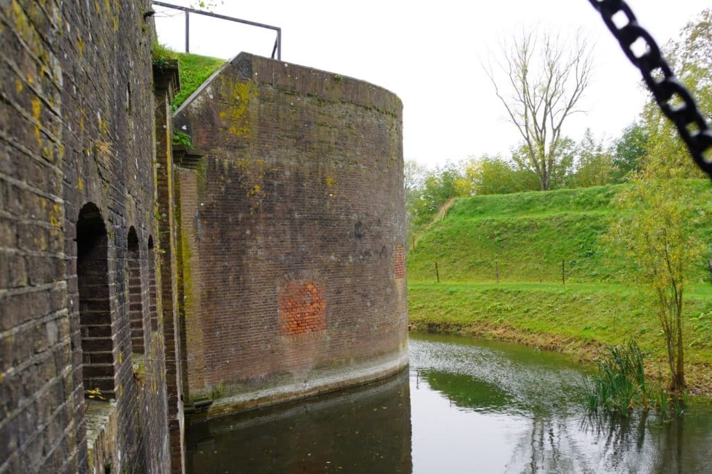 Ein massiver, runder Backsteinturm der Utrechter Wasserlinie ragt direkt aus einem ruhigen Wassergraben empor, flankiert von einer Festungsmauer mit Schießscharten und grünen Graswällen im Hintergrund.