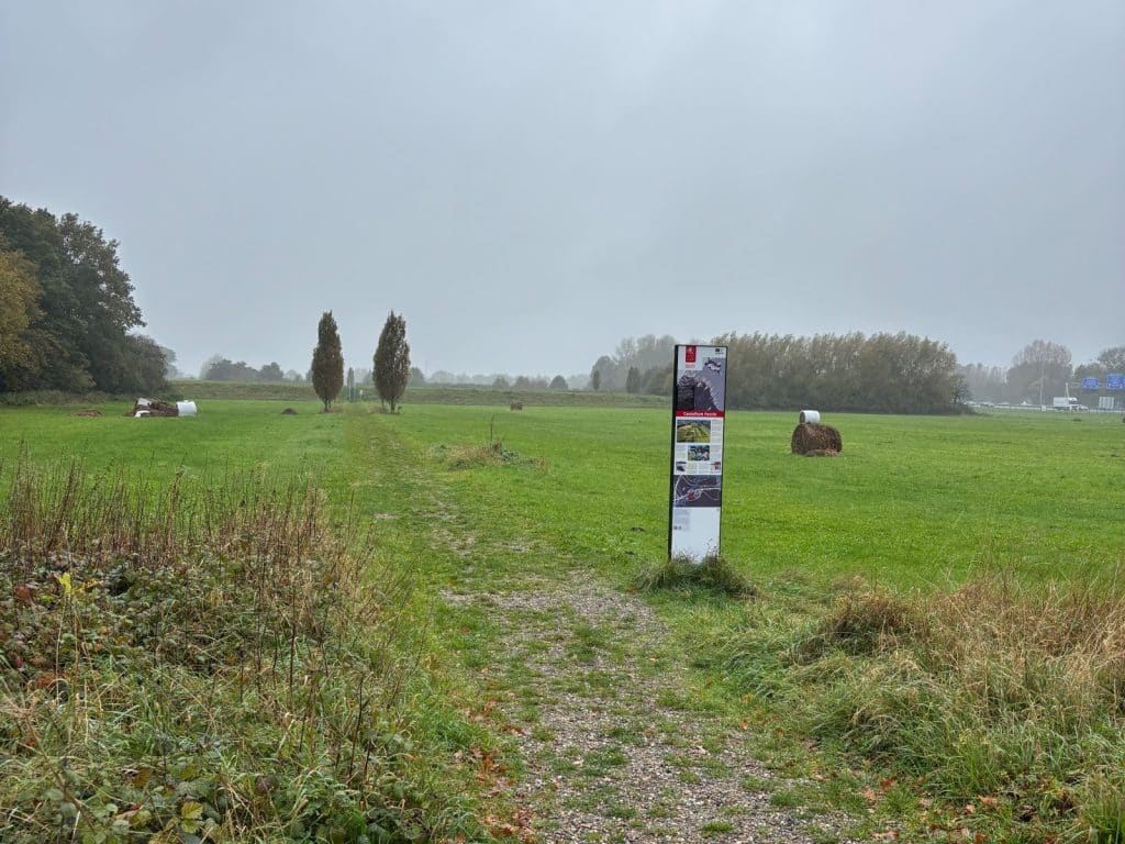 Ein schmaler Pfad führt über eine grüne Wiese am Castellum Fectio bei Utrecht, vorbei an einer Informationstafel der Neuen Holländischen Wasserlinie und zwei markanten, schmalen Bäumen unter einem grauen Himmel.