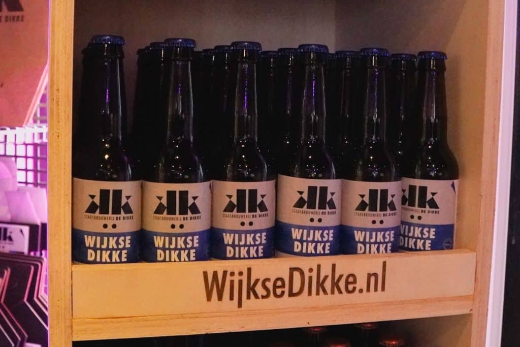 In einem Holzregal stehen mehrere Reihen dunkler Bierflaschen der Brauerei Stadsbrouwerij De Dikke aus Wijk bij Duurstede mit der Aufschrift „Wijkse Dikke“.
