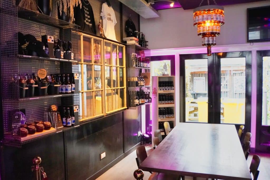 Der moderne Tastingraum der Brauerei in Wijk bij Duurstede zeigt einen langen Holztisch unter einem Kronleuchter aus Bierflaschen, flankiert von Regalen mit Merchandise und Flaschen sowie einer Fensterfront zum Außenbereich.